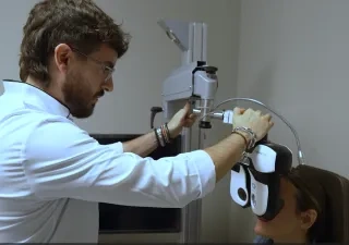 Profesional sanitario realiza una prueba de visión a una persona sentada utilizando un equipo optométrico en una consulta. La persona examinada lleva colocado un dispositivo frente a los ojos mientras el especialista ajusta el aparato para evaluar su capacidad visual. El entorno es una sala clínica con equipamiento oftalmológico.