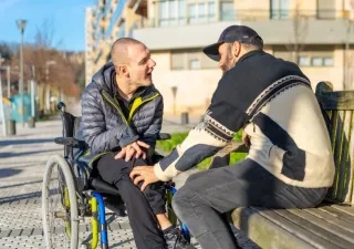 Dos hombres, un de ellos en silla de ruedas y el otro sentado en un banco, hablando
