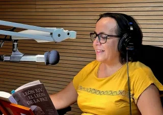 Persona con auriculares sentada en un estudio de grabación frente a un micrófono profesional mientras lee el libro León XIV, el León de la paz, de Almudena Hernández. La imagen muestra la grabación de la versión sonora de la obra, con paneles acústicos de madera al fondo y el ejemplar abierto sobre un atril.