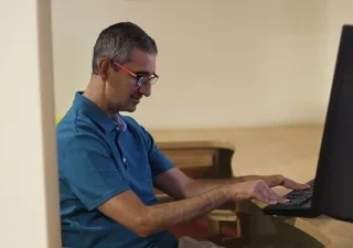 Persona sentada frente a un ordenador de sobremesa, utilizando el teclado en un entorno de trabajo interior. La imagen muestra un puesto de oficina con una pantalla, una mesa de madera y una iluminación suave.