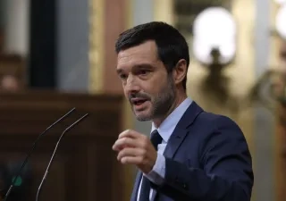 Persona con traje oscuro interviene desde un atril con dos micrófonos en el interior de una cámara parlamentaria. La imagen muestra a la persona realizando un gesto con la mano mientras se dirige al público, con el fondo desenfocado y elementos arquitectónicos de la sala visibles.