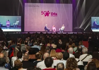 Dos personas subidas en el escenario hablando sentados en dos sillas, mientras hay multitud de personas en la grada observando 