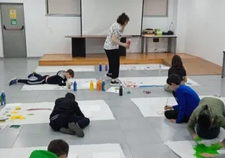 Alt text:  Grupo de menores participa en una actividad artística inclusiva en una sala escolar, pintando sobre grandes hojas de papel colocadas en el suelo. Varias personas trabajan con pinceles y pintura de colores mientras una profesional supervisa la actividad. La imagen refleja un entorno educativo colaborativo orientado al aprendizaje y la participación.
