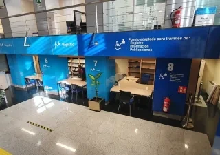 Vista interior de la Oficina de Información y Registro del Paseo del Prado, con varios puestos numerados (6, 7 y 8) señalizados con grandes paneles azules. Cada puesto incluye pictogramas, colores diferenciados y símbolos de accesibilidad. Se observan mesas, sillas, estanterías y un ascensor cercano. La señalética es clara y de alto contraste para facilitar la orientación.