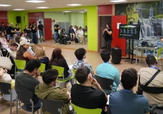 Grupo de personas asistiendo a una sesión formativa o conferencia en una sala accesible. Una persona realiza una presentación frente al público junto a una pantalla, mientras otra persona interpreta en lengua de signos. Entre los asistentes hay personas usuarias de silla de ruedas. La sala cuenta con paredes de colores vivos y un mural de cascadas al fondo.