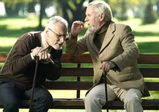 Dos hombres mayores sentados en un parque, acercándose el uno al otro para oirse al hablar