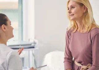 Persona sentada en una consulta médica conversa con un profesional sanitario. Ambos aparecen en una sala de exploración luminosa, con equipamiento clínico al fondo, en una escena relacionada con el asesoramiento y seguimiento de la salud durante la menopausia.