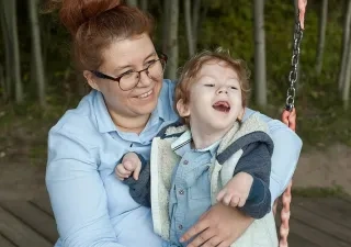 Persona adulta con gafas sostiene a un niño con discapacidad en un columpio adaptado al aire libre. Ambos se encuentran en una zona de juego rodeada de árboles. La imagen refleja acompañamiento, inclusión y apoyo a la infancia con discapacidad en un entorno accesible y seguro.