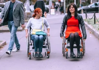 Dos personas usuarias de silla de ruedas se desplazan por una vía urbana peatonal. Una de ellas viste una camiseta clara y lleva un bolso cruzado, mientras que la otra viste un vestido rojo y una chaqueta oscura. A su alrededor caminan otras personas y se observan vehículos aparcados, árboles y mobiliario urbano al fondo. La escena transcurre al aire libre en un entorno urbano accesible.