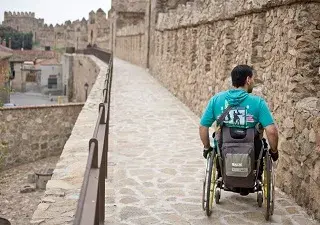 Una persona en silla de ruedas de espaldas transitando por las calles y la muralla de Ávila (Fuente: Ciudades Patrimonio)