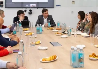 Varios representantes sentados en una mesa hablando, se observan papeles, botellas de agua y comida en el centro de la mesa