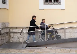hombre ayudando a una persona en silla de ruedas subir por una rampa accediendo a un edificio.