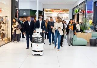 Persona guiando un robot asistencial autónomo por el pasillo de un centro comercial. Varias personas acompañan la demostración y observan el recorrido del dispositivo, en un espacio comercial amplio y luminoso con tiendas y zonas de descanso al fondo. La imagen muestra una prueba de tecnología orientada a mejorar la accesibilidad y la movilidad en entornos comerciales.