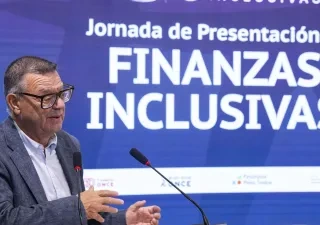 Alt text:  Persona interviniendo desde un atril durante la jornada de presentación de “Finanzas Inclusivas”. La imagen muestra a la persona junto a dos micrófonos mientras realiza una exposición ante el público. Al fondo aparece una gran pantalla azul con el texto “Jornada de Presentación de Finanzas Inclusivas” y los logotipos de las entidades organizadoras. El acto forma parte de la presentación de la renovación del recurso formativo impulsado por Fundación ONCE para promover la educación financiera inclu
