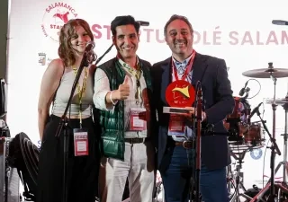 Tres personas posan sobre un escenario durante un acto de Startup Olé Salamanca. Una de ellas sostiene un premio de color rojo mientras otra hace el gesto de aprobación con el pulgar. En primer plano hay varios micrófonos y, al fondo, se aprecia una batería y el logotipo del evento proyectado en una pantalla.