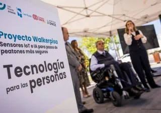 Presentación del proyecto WalkerPisa de Fundación ONCE en un paso de peatones de Las Rozas. En primer plano, un cartel con el lema “Proyecto Walkerpisa. Sensores IoT e IA para pasos de peatones más seguros. Tecnología para las personas”. Al fondo, varias personas durante el acto de presentación, una de ellas utilizando una silla de ruedas, en un entorno urbano al aire libre.