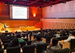 Auditorio moderno con varias decenas de personas sentadas en butacas, asistiendo a una conferencia. En el escenario, una persona realiza una presentación desde un atril mientras otra permanece sentada en una mesa de ponentes. Al fondo se proyecta una diapositiva titulada “Consecuencias del delirium”. La sala cuenta con iluminación cálida, paredes de madera y una amplia zona de gradas ocupada por el público.