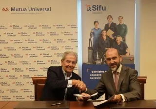 Dos representantes de Mutua Universal y Grupo SIFU estrechan la mano durante la firma de un acuerdo de colaboración. Ambos están sentados frente a una mesa con documentación, junto a paneles corporativos de las dos entidades relacionados con la integración laboral y la inclusión de personas con discapacidad.