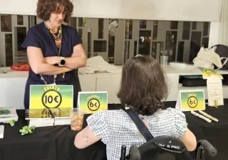 Una persona en silla de ruedas de espaldas atendiendo en el mercadillo a una señora que viene a comprar