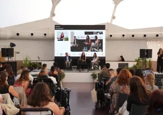 Participantes asisten a una mesa redonda del II Congreso Estatal de Juventud con Discapacidad en Valladolid. En el escenario, tres ponentes intervienen en un debate sobre los 20 años de la Convención de la ONU sobre los Derechos de las Personas con Discapacidad, mientras una intérprete de lengua de signos presta apoyo comunicativo. Al fondo, una gran pantalla reproduce la sesión y, entre el público, se observan asistentes con y sin discapacidad siguiendo la jornada.