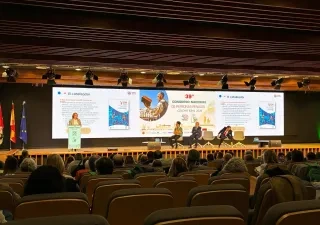 Vista general de un auditorio durante una ponencia del 39º Congreso Nacional de Personas Renales, celebrado en Ciudad Real en 2026. En el escenario, una persona interviene desde un atril mientras varias personas permanecen sentadas en una mesa redonda. Al fondo, una gran pantalla proyecta una presentación sobre el espacio GN Conecta y el papel de “El catalizador”. El público ocupa gran parte de las butacas del auditorio, orientadas hacia el escenario iluminado. A la izquierda se observan las banderas de Esp