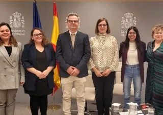 Varias personas posan de pie en una sala institucional frente a un fondo gris con el logotipo del Gobierno de España y dos banderas, una de la Unión Europea y otra de España. Delante hay una mesa con documentos, micrófonos y vasos de agua. El grupo está alineado en el centro del espacio, con sillas blancas en los laterales.