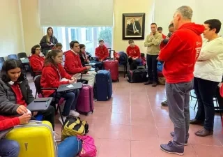 Grupo de jóvenes con sudaderas rojas sentados en un aula o sala de reuniones junto a varias maletas de viaje de distintos colores. En la parte frontal de la sala, varias personas ofrecen indicaciones antes de una salida o actividad organizada. El espacio cuenta con sillas, pupitres, grandes ventanales y equipaje distribuido por la estancia, en un ambiente de preparación para un desplazamiento colectivo.