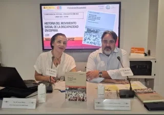Mesa de presentación de una publicación del CERMI sobre la historia del movimiento social de la discapacidad en España. Dos personas participan en una conferencia junto a ejemplares del libro, micrófonos y material de trabajo. Al fondo, una pantalla muestra la portada de la obra y el título de la presentación virtual.
