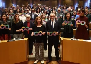salón de actos del Congreso de los Diputados, lleno de asistentes situados tanto de pie como sentados. Todas las personas sostienen carteles con el símbolo del infinito multicolor, representativo de la diversidad del espectro del autismo. En primera fila, varios participantes permanecen de pie junto a los escaños, mientras el resto del público ocupa el fondo del auditorio. El ambiente es institucional y de apoyo colectivo a la causa del autismo.