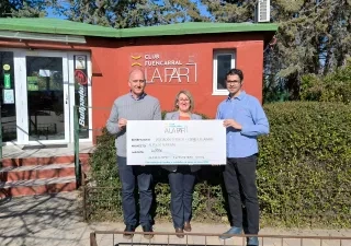 Tres personas posan frente a la sede del Club Fuencarral A LA PAR sosteniendo un cheque simbólico de ayuda económica para un proyecto deportivo dirigido a personas con discapacidad intelectual. Al fondo se ve el edificio del club y su rótulo identificativo.