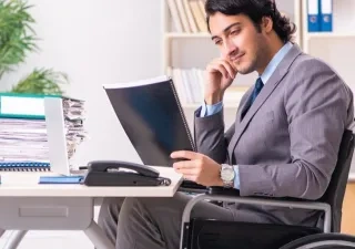 Persona con discapacidad física que va en silla de ruedas traajando en el escritorio con su ordenador y cuadernos.