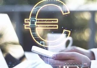 Mano sosteniendo un teléfono móvil junto a un ordenador portátil, con un símbolo del euro digital superpuesto en el centro de la imagen. La escena representa conceptos relacionados con pagos electrónicos, finanzas digitales y tecnología financiera. Fondo desenfocado con tonos claros y elementos tecnológicos.