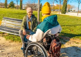 Persona en silla de ruedas y otra persona mantienen una conversación en un parque urbano con zonas verdes y caminos peatonales.
