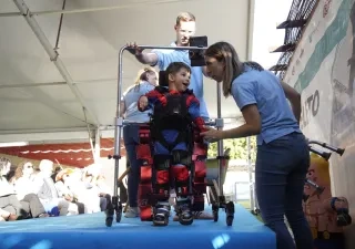 Niño utilizando un exoesqueleto pediátrico asistido por profesionales durante una demostración de neurorrehabilitación. El menor se encuentra sujeto a un dispositivo robótico de marcha sobre una plataforma, mientras personal especializado supervisa y ajusta el equipo ante un público asistente.