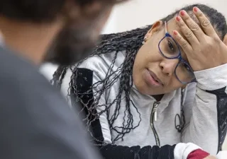 Persona con gafas conversa con otra persona en un entorno interior. Apoya la cabeza sobre una mano mientras escucha, en una escena relacionada con atención, apoyo o acompañamiento profesional.