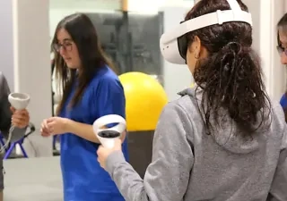 Persona utilizando unas gafas de realidad virtual y mandos de control durante una actividad de rehabilitación tecnológica en un centro especializado. En la sala hay otras dos profesionales con uniforme azul y una pelota terapéutica amarilla al fondo.