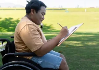 Niño en silla de ruedas escribiendo en un cuaderno a la sombra en un césped
