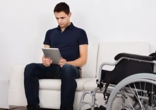 Persona con movilidad reducida sentado en el sillón junto a su silla de ruedas, mientras usa la tablet