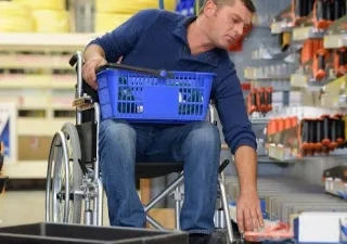 Interior de una tienda de bricolaje o ferretería. Una persona en silla de ruedas se desplaza por un pasillo comercial mientras sostiene una cesta de compra azul sobre las piernas. Con una mano alcanza un producto situado en un estante inferior. A ambos lados del pasillo hay estanterías con herramientas, accesorios y materiales organizados. La imagen muestra una situación de compra y accesibilidad en un entorno comercial.