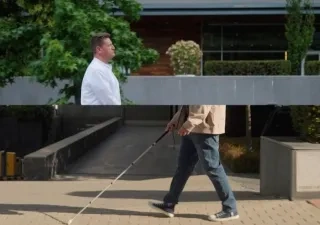 Dos personas aparecen en un entorno al aire libre con un edificio moderno y vegetación de fondo. La persona de la izquierda, vestida con una camisa blanca, está de pie cerca de un seto y árboles. La persona de la derecha, con una chaqueta beige, jeans azules y zapatillas, camina con un bastón blanco, navegando por un sendero pavimentado con una rampa y un muro bajo