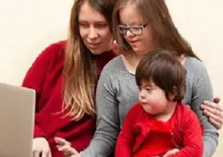 Una persona enseñando a manejar un ordenador a otras dos con discapacidad cognitiva (una adolescente y un bebé)