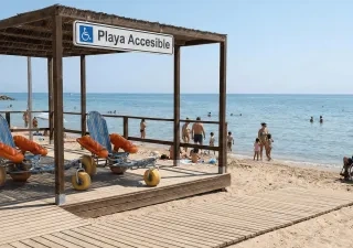 **Alt text:**  Playa con servicio accesible señalizado (“Playa Accesible”) bajo una estructura de madera con pasarela hasta la orilla, donde hay varias sillas anfibias con flotadores naranjas; al fondo, personas en la arena y baño en el mar, y a la derecha una persona usuaria de silla de ruedas acompañada junto al agua.