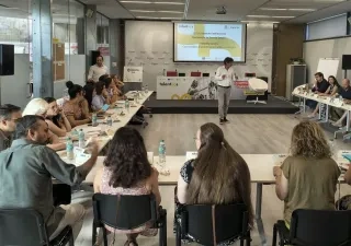 Muchas personas sentadas en una mesa con una ponente en el medio que modera y dirige la sesión