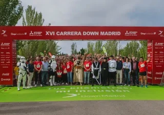 Participantes de la XVI Carrera Down Madrid posan bajo el arco de salida en el Parque Juan Carlos I antes del inicio de la prueba. En primer plano aparecen corredores, voluntarios y personas disfrazadas de personajes de ciencia ficción, mientras una gran multitud se concentra detrás. El arco rojo muestra el nombre del evento y los logotipos de las entidades colaboradoras. La carrera tiene como objetivo promover la inclusión de las personas con discapacidad intelectual a través del deporte.